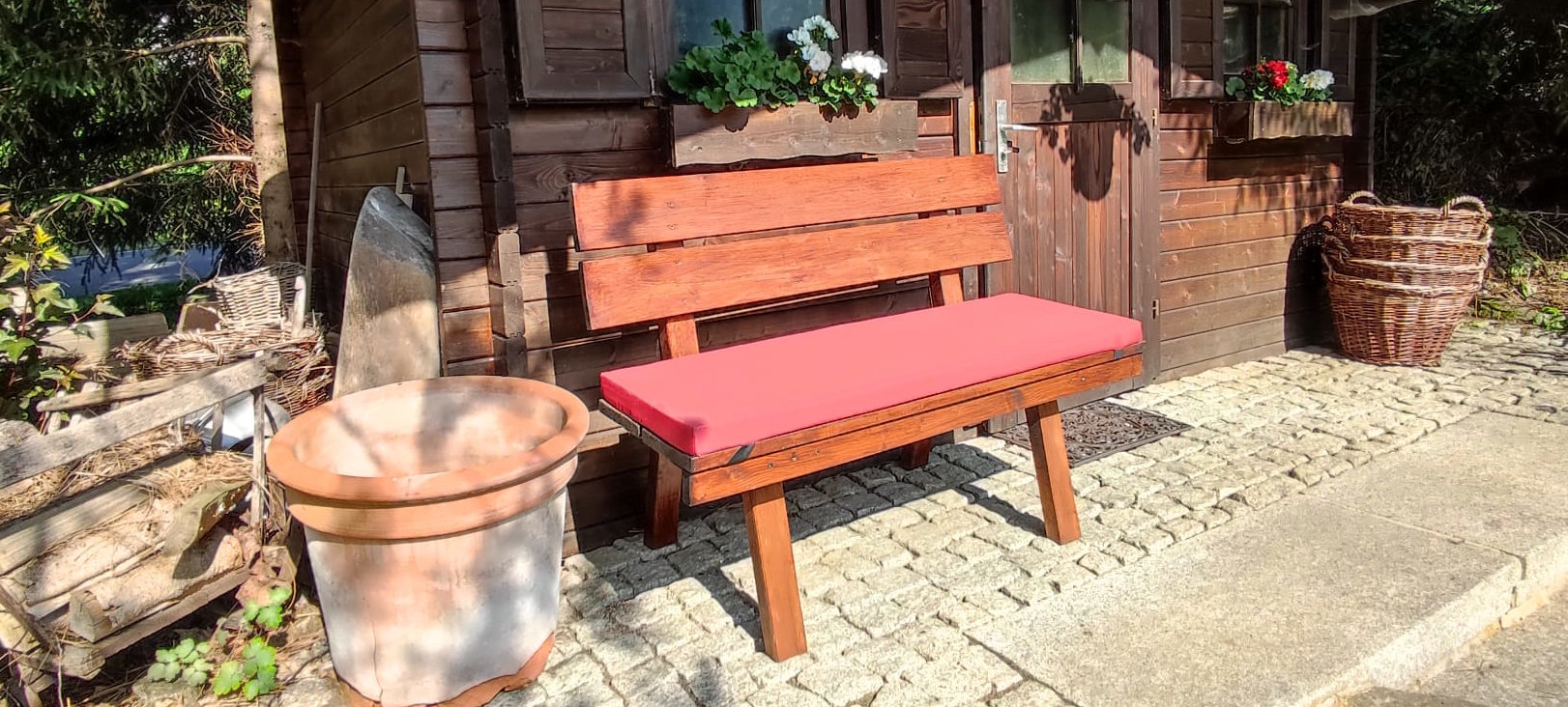 kleine Bank mit rotem Sitzkissen, an Gartenhütte stehend